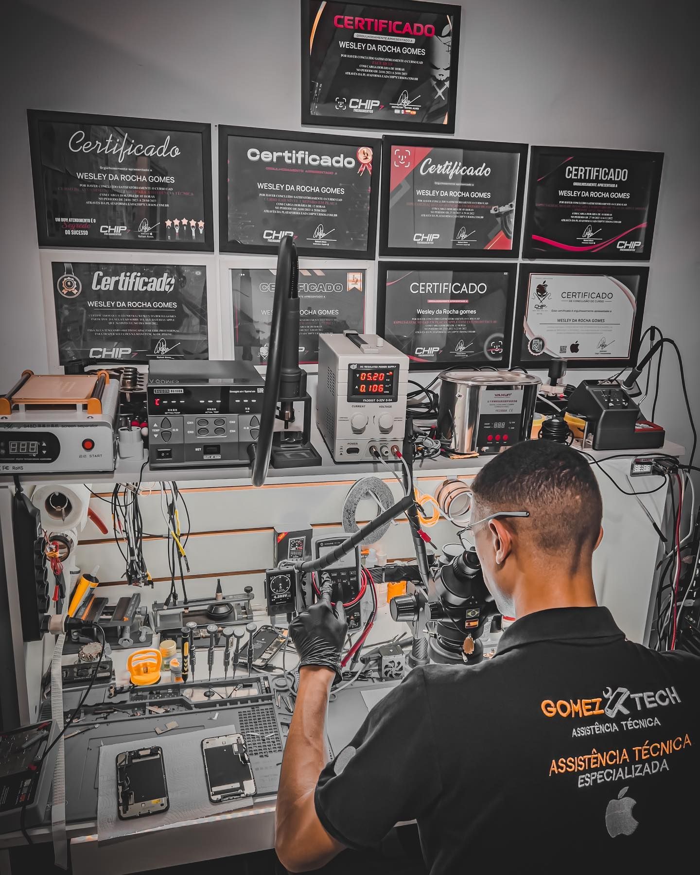 Wesley da Rocha Gomes trabalhando na bancada da Gomez.Tech, com microscópio e equipamentos de reparo; ao fundo, a parede com seus certificados de formação técnica.